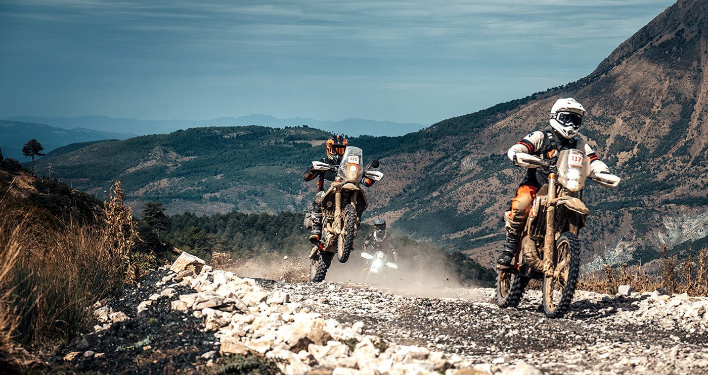 Two riders on a loose rocky mountain track with peaks behind them during Albania Raid